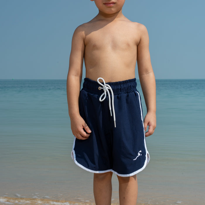 Young boy in navy swim shorts standing on a beach with ocean in the background