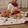 Child sitting on a wooden floor with teddy bears and a blanket, wearing a bib.