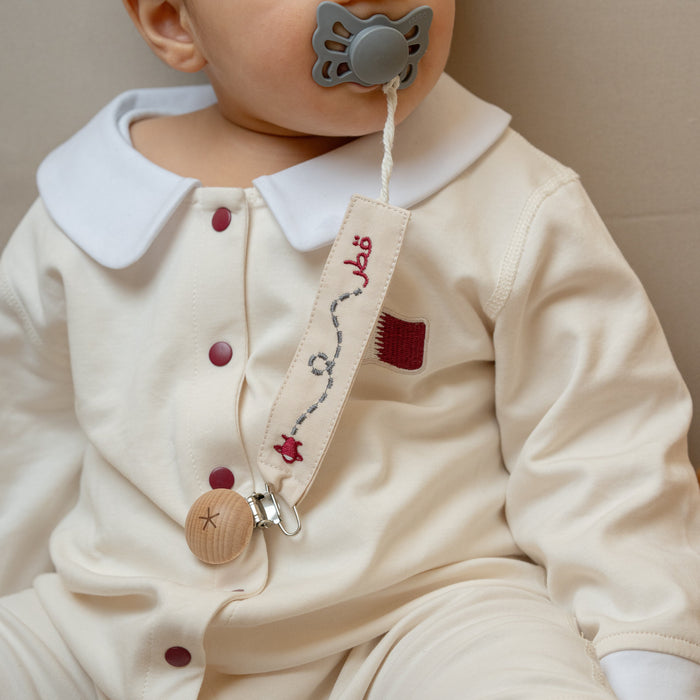 Baby wearing a white outfit with a collar and buttons, holding a pacifier with a strap pacifier holder.