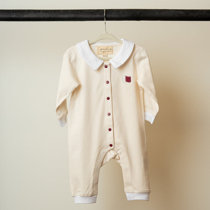 Beige baby romper with white collar and red buttons with embroidered Qatar flag on a hanger against a plain background