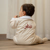Child wearing a baby romper or baby onesie with Arabic Qatar  text sitting on the floor in front of a curtain.