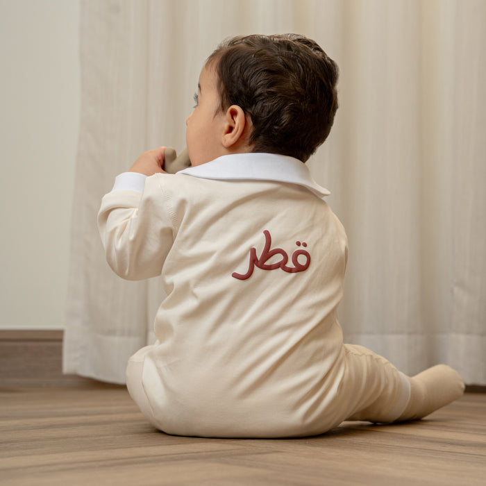 Child wearing a baby romper or baby onesie with Arabic Qatar  text sitting on the floor in front of a curtain.