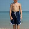 Young boy in navy swim shorts standing on a beach with ocean in the background