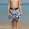 Young boy in coral patterned swim shorts standing on a beach with clear blue water and sky.
