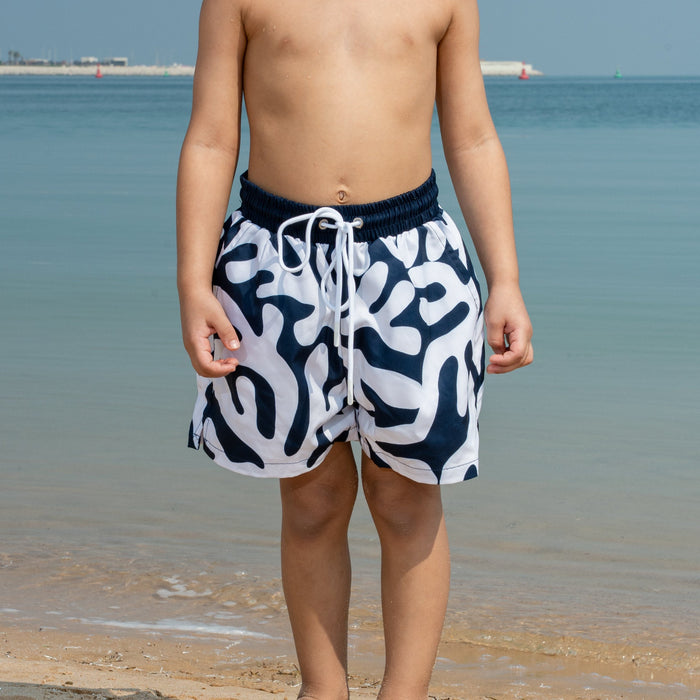 Young boy in coral patterned swim shorts standing on a beach with clear blue water and sky.