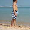 Child in coral-patterned swim shorts standing on a beach with clear blue water and sky.