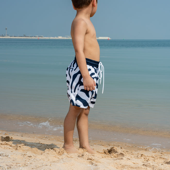 Child in coral-patterned swim shorts standing on a beach with clear blue water and sky.