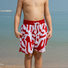Young boy wearing red and white swim shorts standing on a beach.