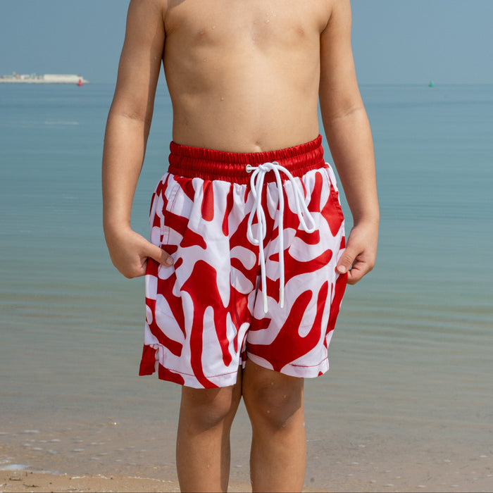 Young boy wearing red and white swim shorts standing on a beach.