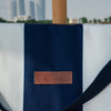 Navy blue and white tote bag with a leather patch on a blurred cityscape background
