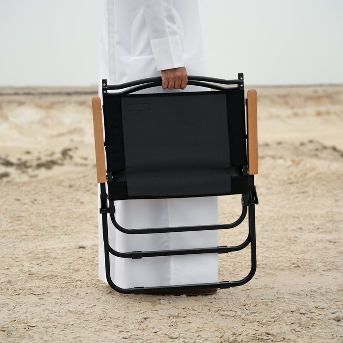 Person holding a black folding chair with wooden armrests on a textured floor.