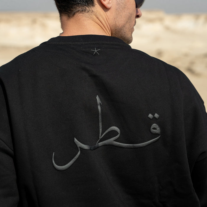 Person wearing a black sweatshirt with Arabic text on the back, standing against a desert landscape.