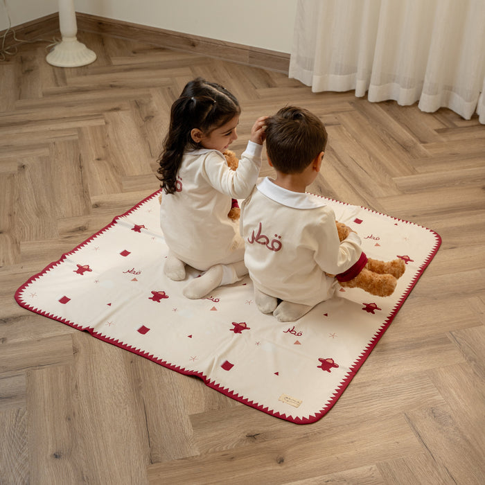 Two children sitting on a Qatar baby blanket with teddy bears in a room with wooden flooring and white curtains.