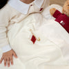 Child wrapped in a white blanket with a teddy bear and Qatar beads safety pins, sitting on a wooden floor.