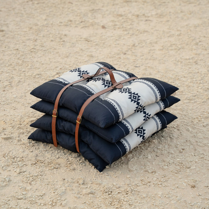 Stack of Sadu patterned pillows or cushions with a brown leather strap on a sandy surface