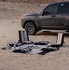 Car parked in a desert with a picnic setup on a Sadu patterned rug, cushions and floor chair