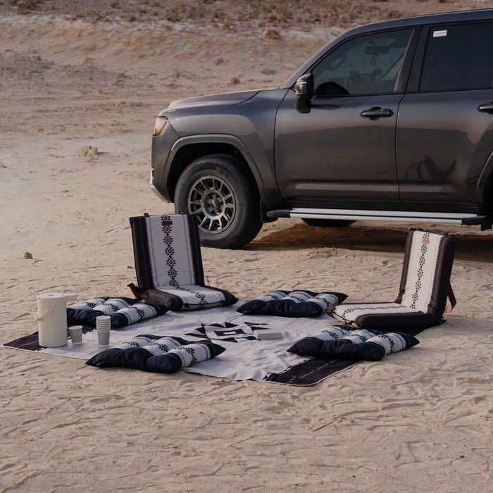 Car parked in a desert with a picnic setup on a Sadu patterned rug, cushions and floor chair