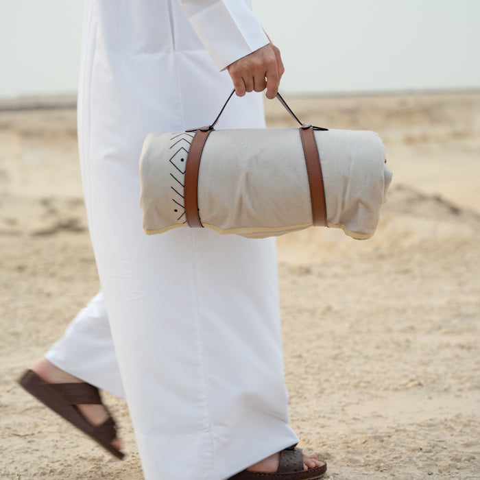Person ina thoub holding a rolled carpet with brown handle on sandy ground