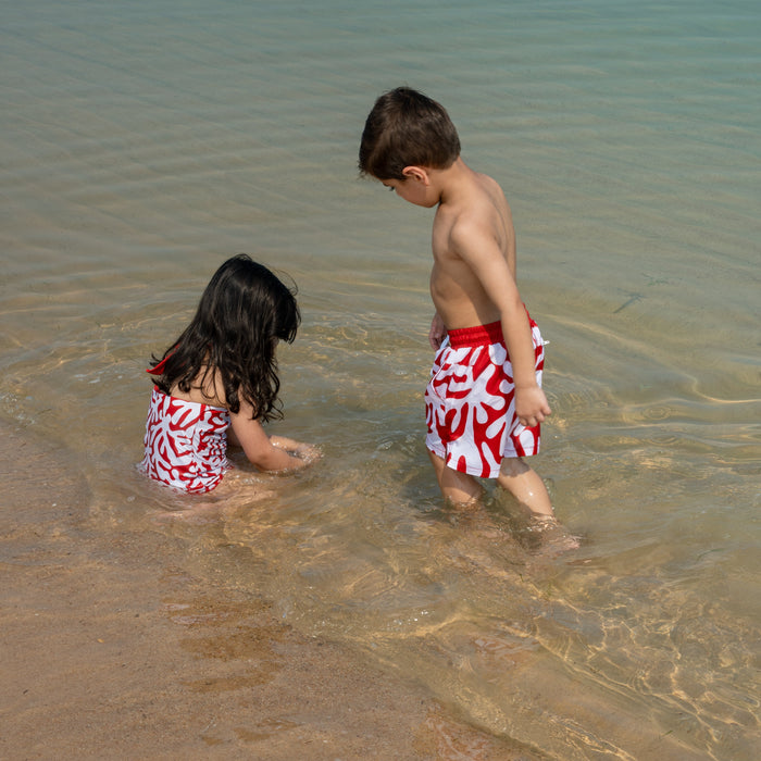Two children in matching red and white  coral patterned swimsuits playing in shallow water at the beach.