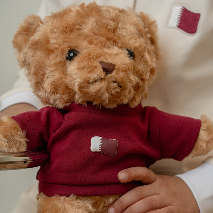 Brown teddy bear wearing a custom t shirt with a Qatar flag print logo, held by a baby.