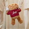 Two children in matching outfits holding a teddy bear with Arabic Qatar name in text, standing on a wooden floor.