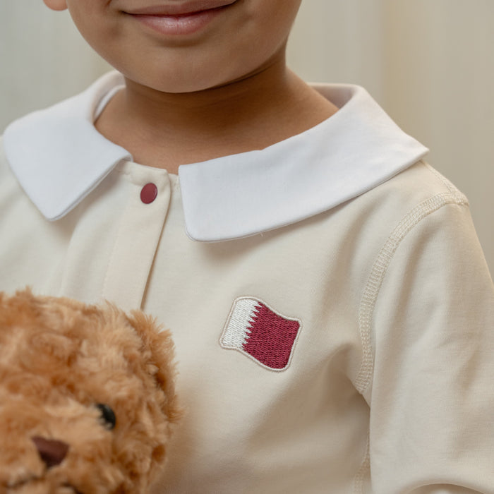 Child wearing a cream-colored shirt with a an embroidered Qatar flag, holding a teddy bear.