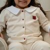 Young girl wearing a beige toddler suit with a white  collar, embroidered Qatar flag  and red button, sitting on a beige chair.