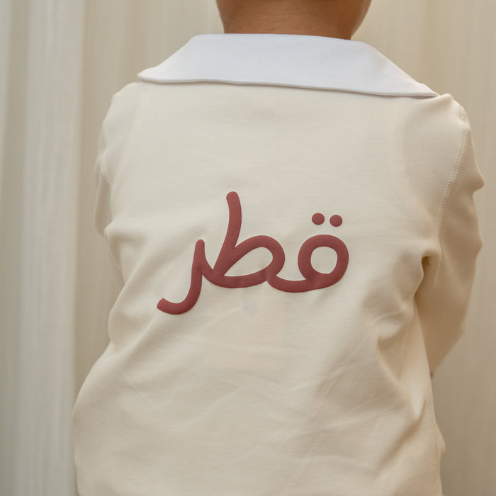 Child wearing a beige toddler suit with Arabic Qatar text on the back against a neutral background